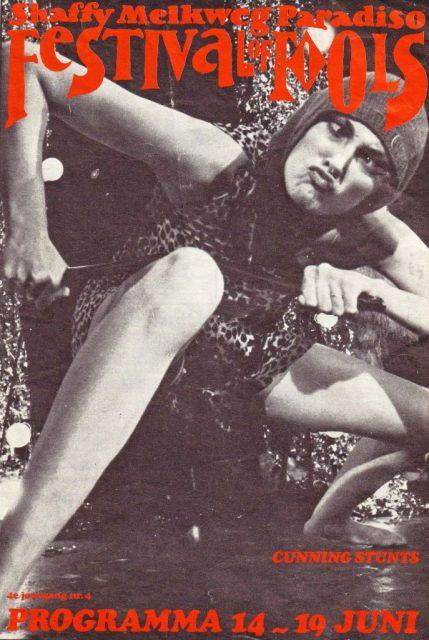 A programme for The Festival of Fools in Amsterdam in 1979. A woman is squatting close to the ground in a leopard print dress, breaking a stick over her leg. She is wearing a balaclava and crossing her eyes and pouting.