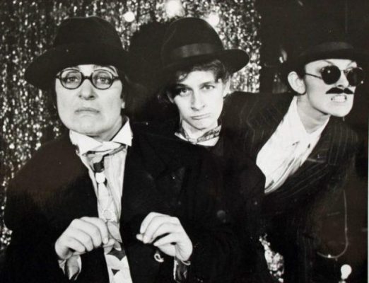 A black and white image of three women in suits and fedoras, two looking directly at the camera, one snarling away from the camera in sunglasses and a fake moustache