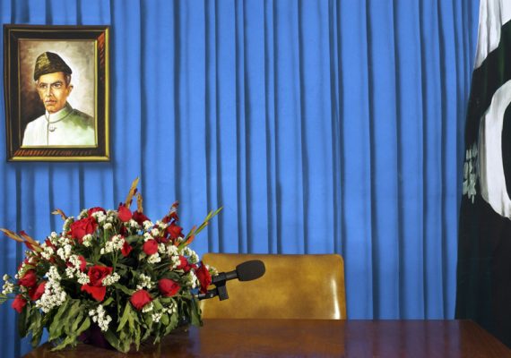 an chair behind an empty table. The background is a pleated blue curtain, and there is a bouquet of roses on the table. a portrait of Muhammad Ali Jinnah hangs on one side, a folded flag of pakistan on the other.