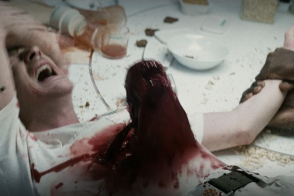 a still from the film Alien, in which a creature is bursting out of a character's chest, covered in blood. The character is visibly in pain and covering their eyes.