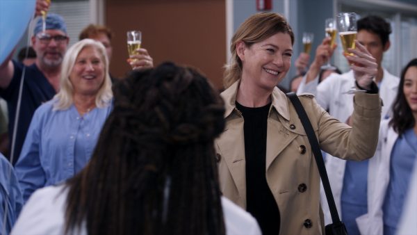 A still from the episode of Grey's Anatomy in which Meredith leaves. She is smiling and raising a glass of champagne wearing a trench coat and a shoulder bag. She is surrounded by colleagues also holding up glasses, everyone else visible is smiling.