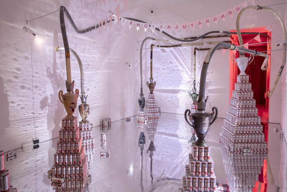 a white room with a mirrored floor, inside which there are pyramids of energy drink cans, topped with rat-face-shaped trophies, all connected to pipes leading to the wall. there is bunting hanging from the ceiling that reads 'congratulations'