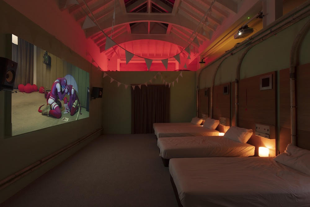 an interior that looks like a motel room, with three beds in a row, the room is dimly lit, there is a video projected onto the wall and there is bunting hanging from the ceiling.